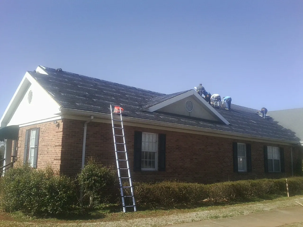Crew tearing shingles off a brick ranch roof with a ladder set — Emergency Roof Repair work by our crew in Alcolu