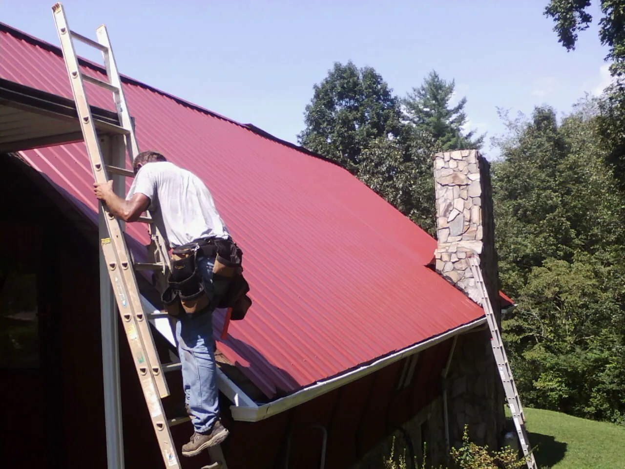 Roofer on a ladder beside a red metal roof and stone chimney — Soffit & Fascia Repair by Roofing Brothers in Alcolu